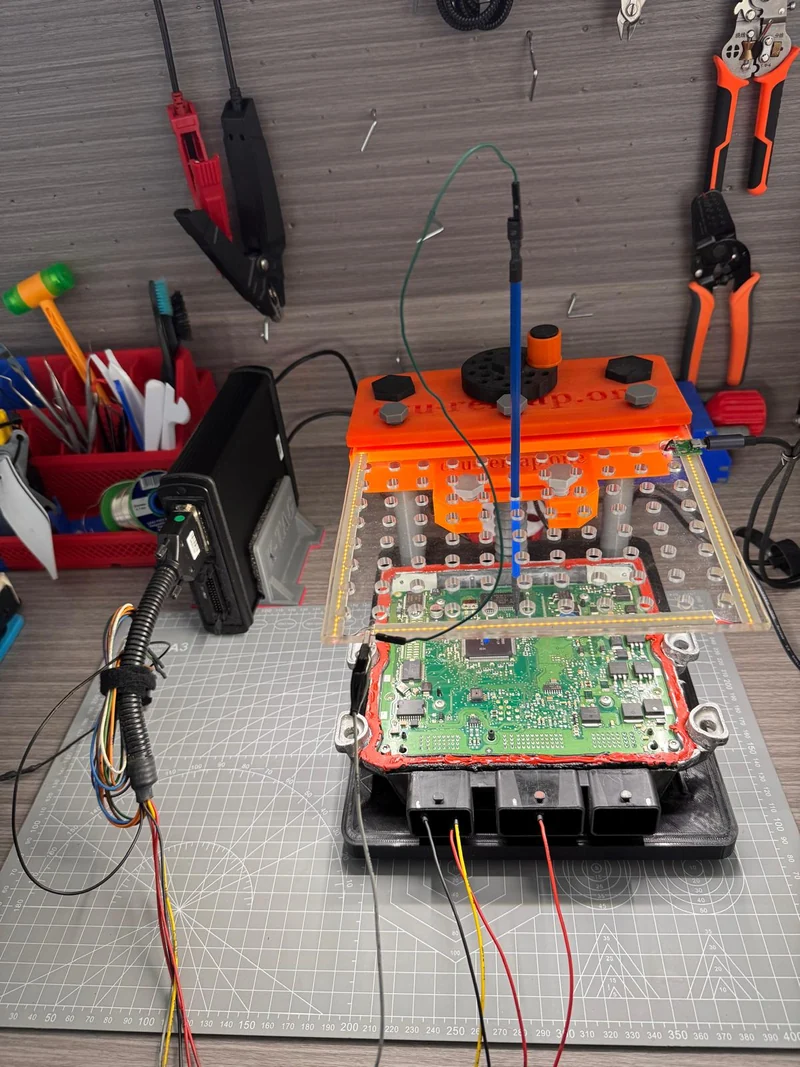 Opened PSA ECU printed circuit board with connected programming probes and orange fixture.